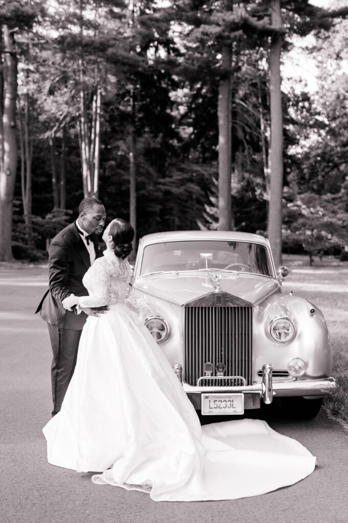 black and white photo of bride and groom next to a vintage car