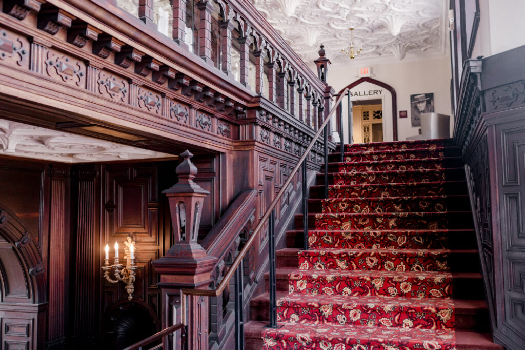 an elegant staircase in Branford House, CT, with a rich red floral carpet, intricate dark wood paneling, ornate railings, and a beautifully detailed ceiling