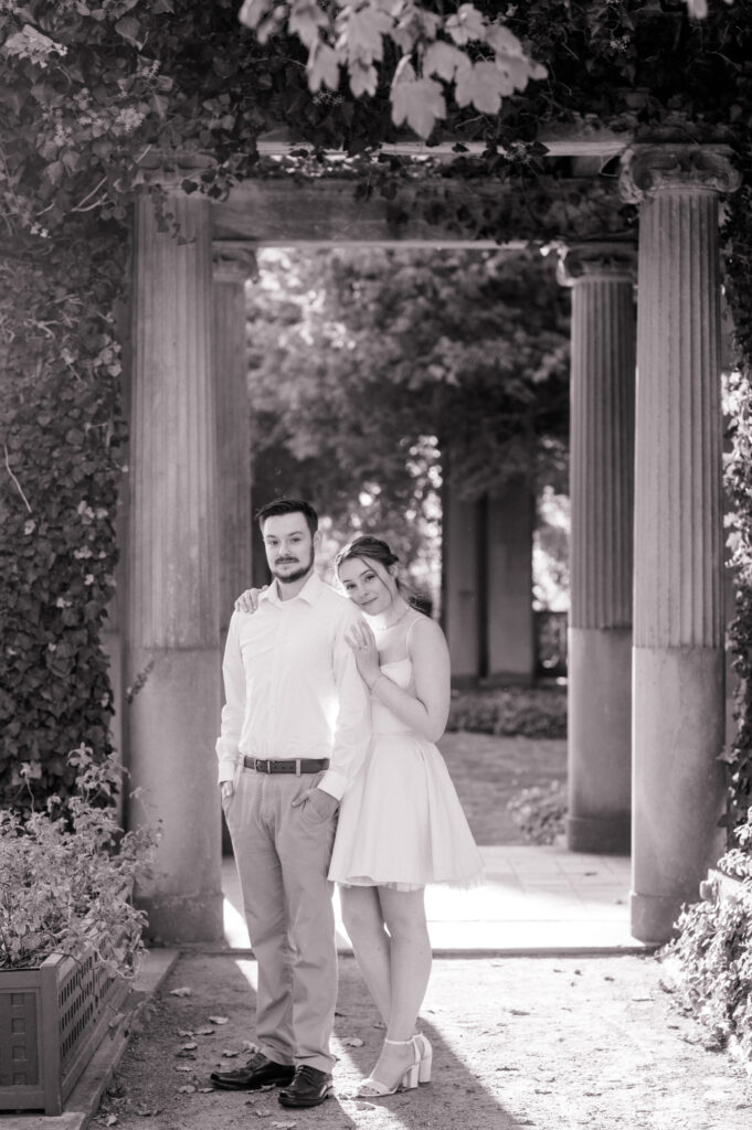 A black-and-white portrait of a bride and groom under a vine-covered archway at Eolia Mansion, surrounded by historic stone columns and soft natural light.