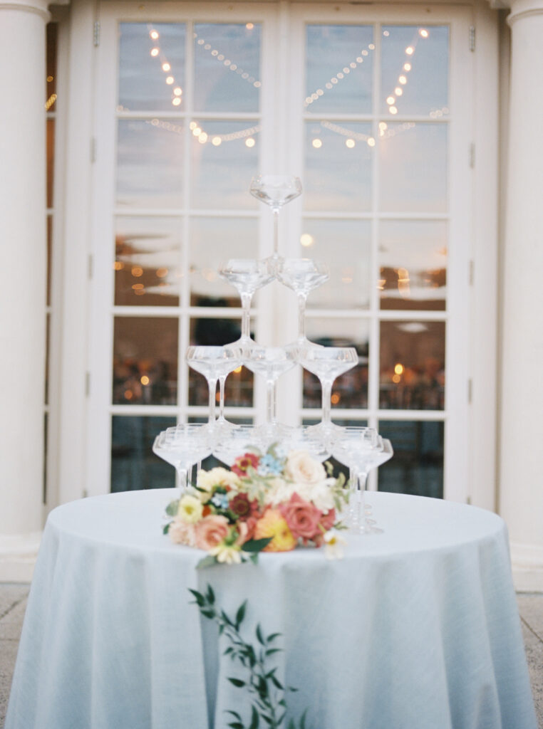 A champagne tower with a floral centerpiece at the base, set against the backdrop of glass doors and string lights, captured on film.