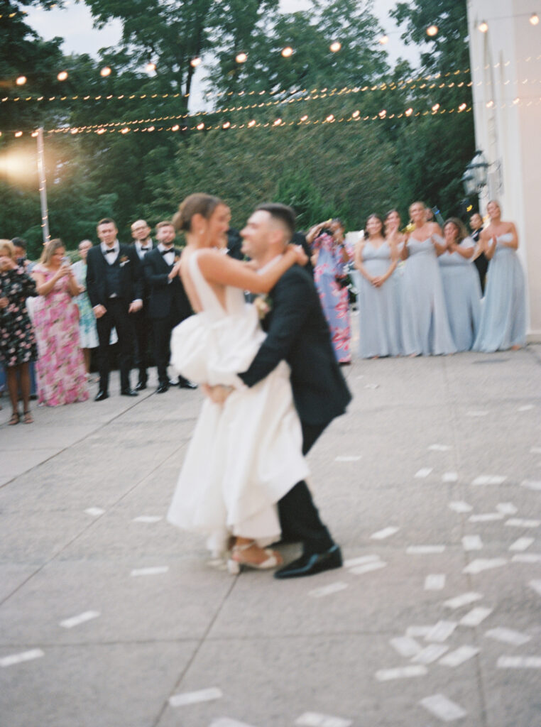 A candid moment of the bride and groom dancing together under twinkling string lights, surrounded by guests, captured on film.