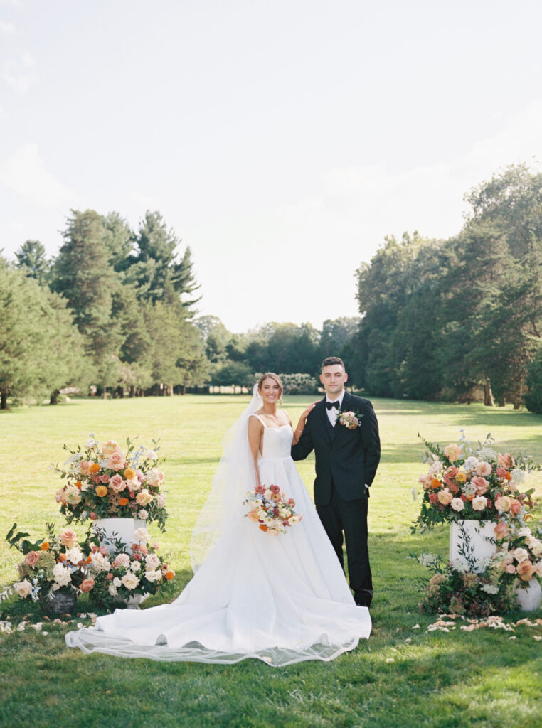 Bride and groom standing together on lawn, framed by beautiful floral arrangements in warm tones, captured on film.