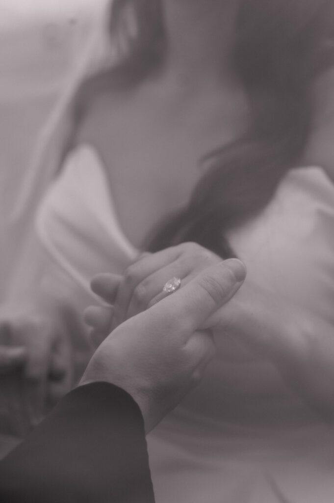 Black and white close up photo of groom holding brides hand, with focus on her engagement ring. 