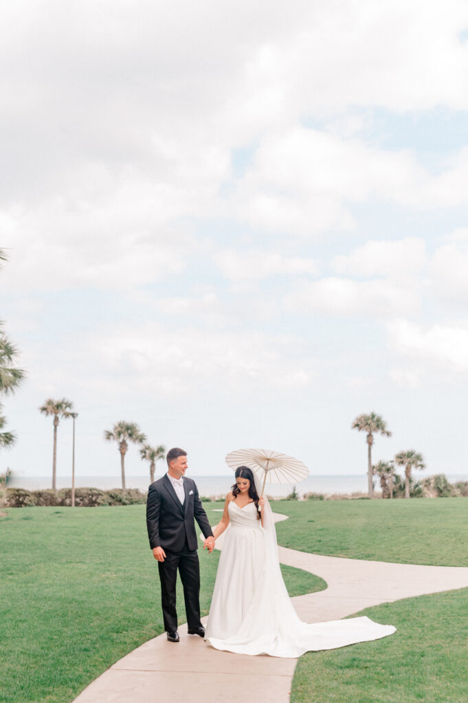 Groom leading bride, walking on sidewalk with beautiful Atlantic ocean and palm trees in the background. 