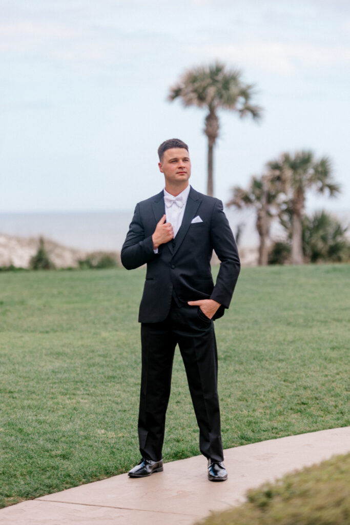 Groom stands on sidewalk getting portraits taken for wedding with the Atlantic ocean and dunes as the backdrop. 