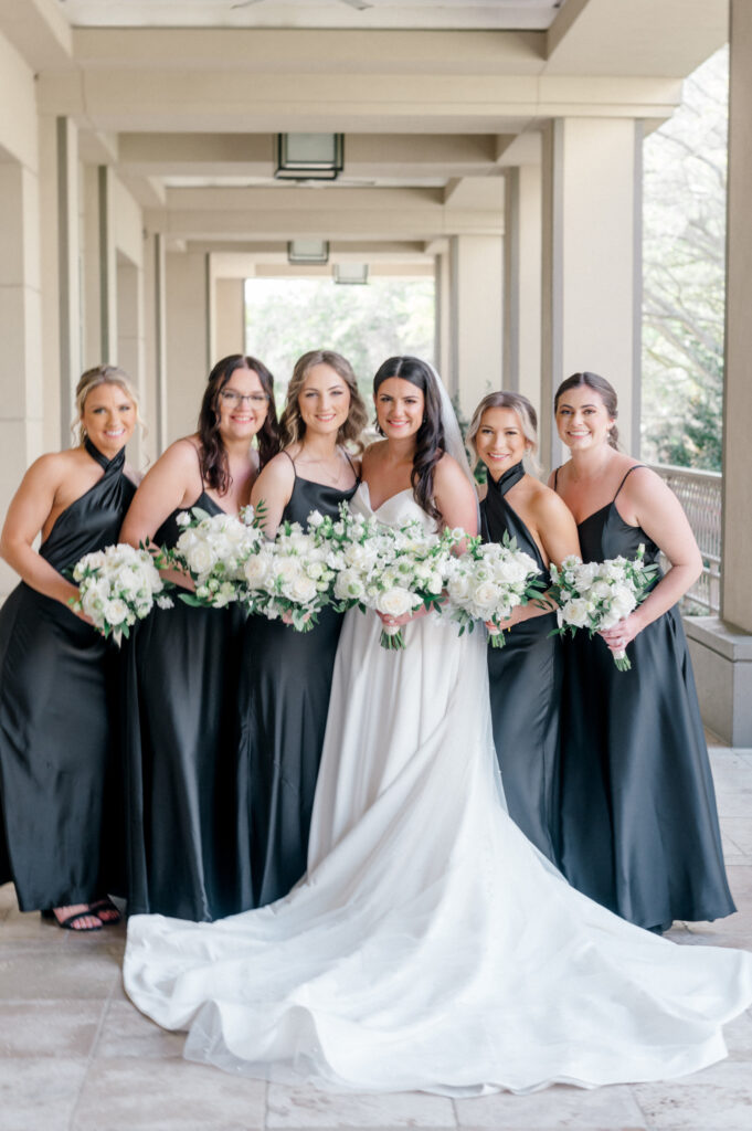 Bride poses with her bridal party wearing all black, satin dresses and holding monochromatic white bouquets with greenery. 