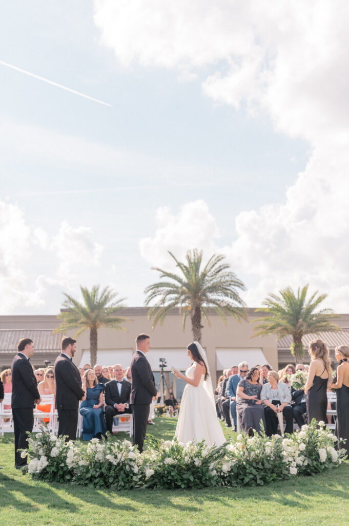 Bride and groom exchange vows during wedding ceremony surrounded by ground-level white floral arch. 