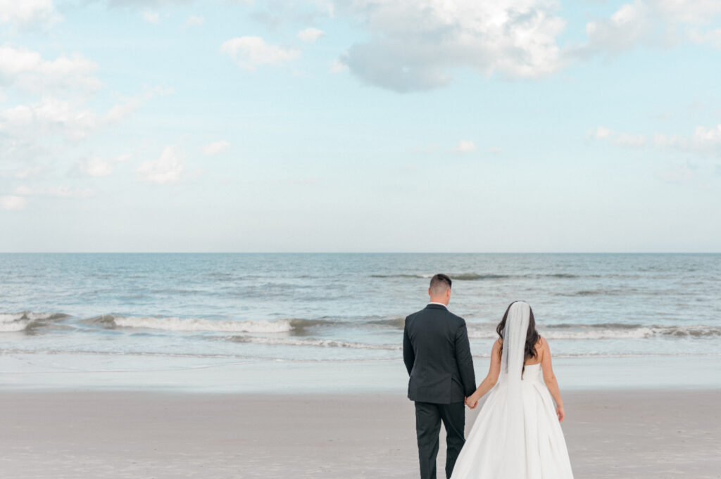 Bride and groom walk together, hand in hand, toward the ocean. 