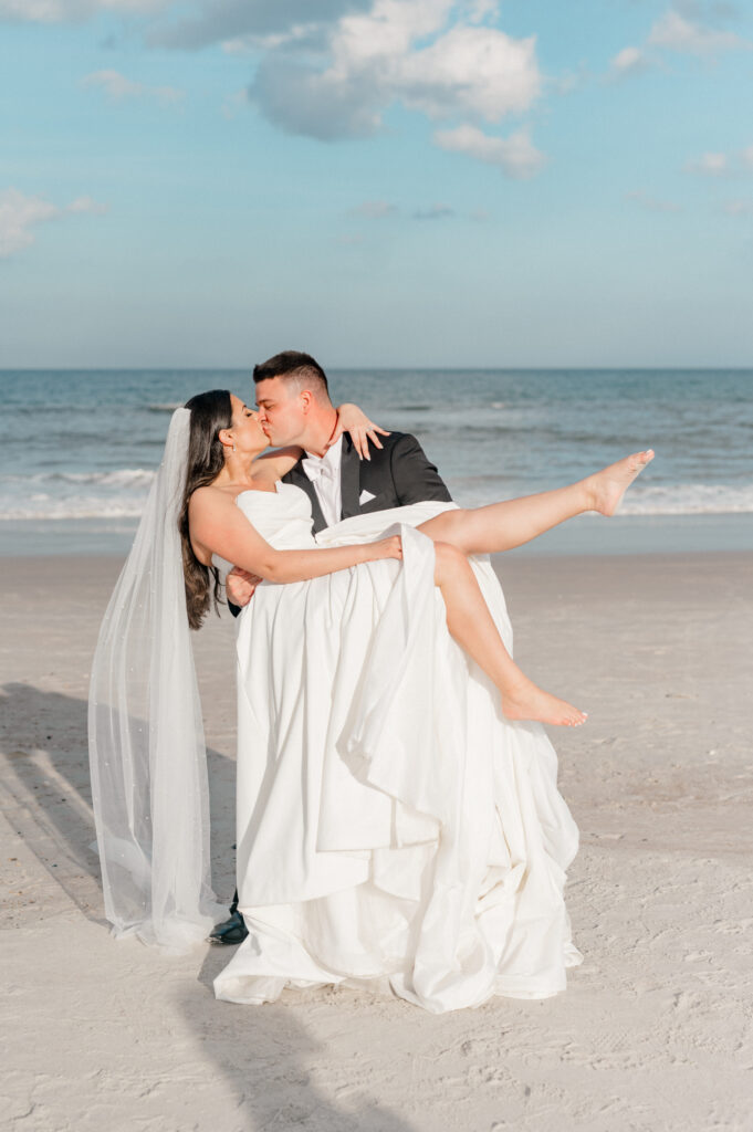 Groom holds bride on beach as they share a kiss for their wedding photography. 