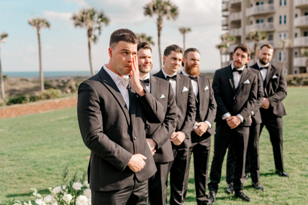 Groom stands at the altar with his groomsmen, holding his face as he cries watching his bride walk down the aisle. 