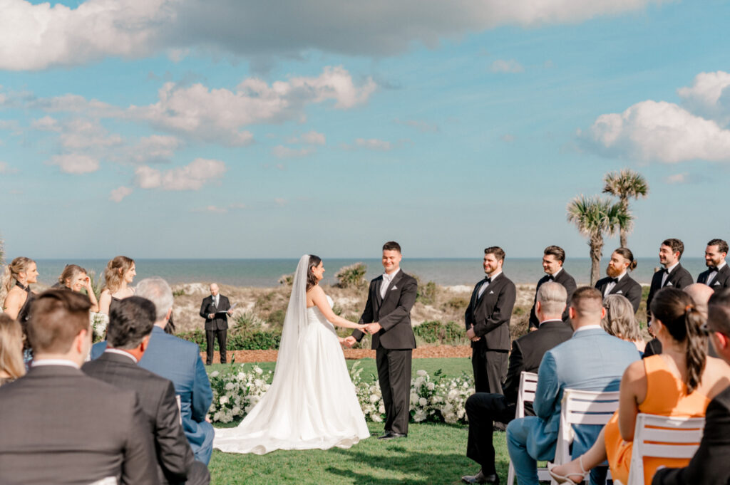 Groom looks into crowd as him and his bride exchange vows during wedding ceremony surrounded by ground-level white floral arch and family. 