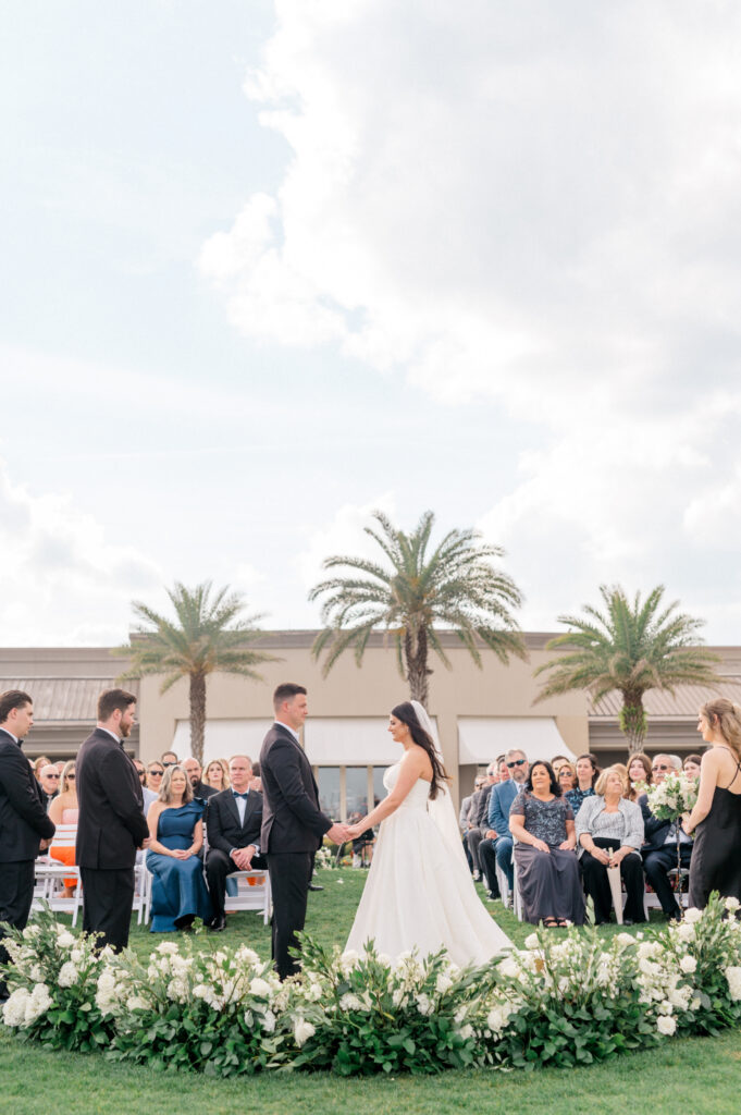 Bride and groom hold hands during wedding ceremony surrounded by ground-level white floral arch. 