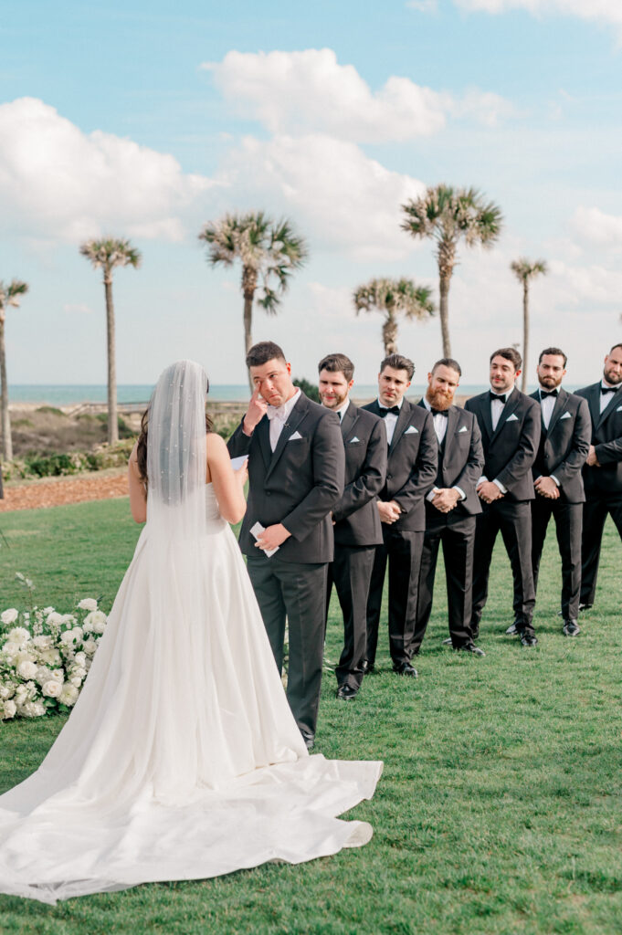 Groom stands at the altar with his groomsmen, holding his face as he cries listening to the vows being read by his bride. 