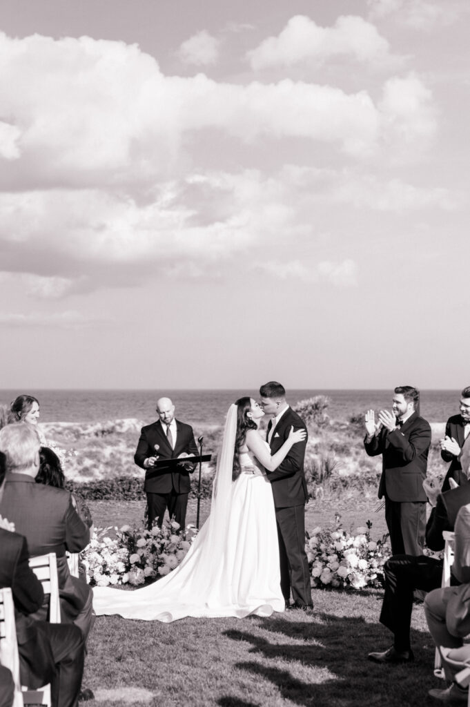 Black and white photo of bride and groom sharing first kiss after wedding at the Ritz Carlton Amelia Island with a stunning view of the dunes and Atlantic ocean behind them. 