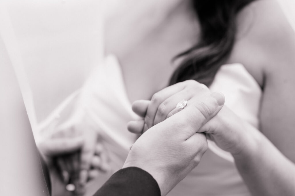 Black and white close up photo of groom holding brides hand, with focus on her engagement ring. 