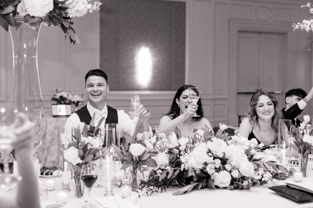  Black and white photo of bride and groom raising a glass after toasts during their wedding reception. 