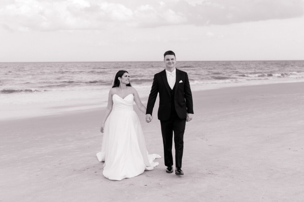 Black and white photo of bride and groom walking together on the beach with the Atlantic ocean behind them. 