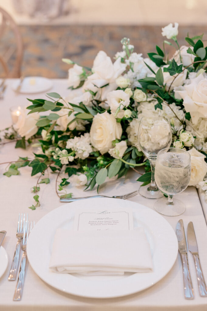 Table setting for wedding reception with white plate and napkin, silver silverware, water glass and white floral arrangement. 