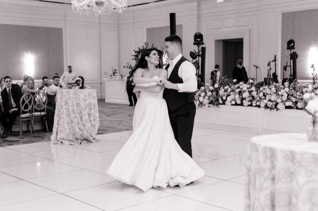 Black and white photo of bride and groom sharing first dance together on white tile dance floor. 