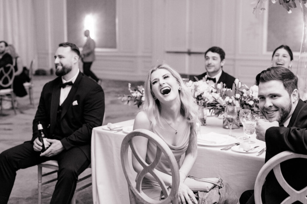 Black and white photo of guests sitting and table and laughing during wedding toasts. 