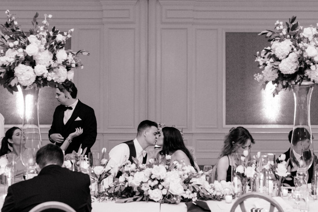  Black and white photo of bride and groom sharing kiss at the head table during their wedding reception. 