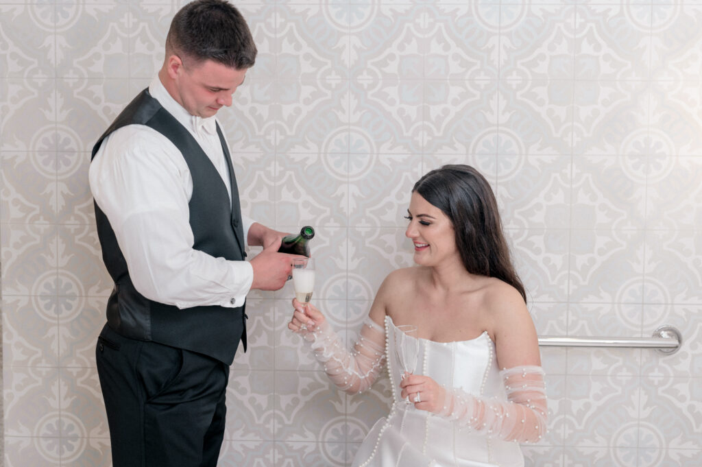 Bride sits holding champagne as groom pours champagne into her glass. 