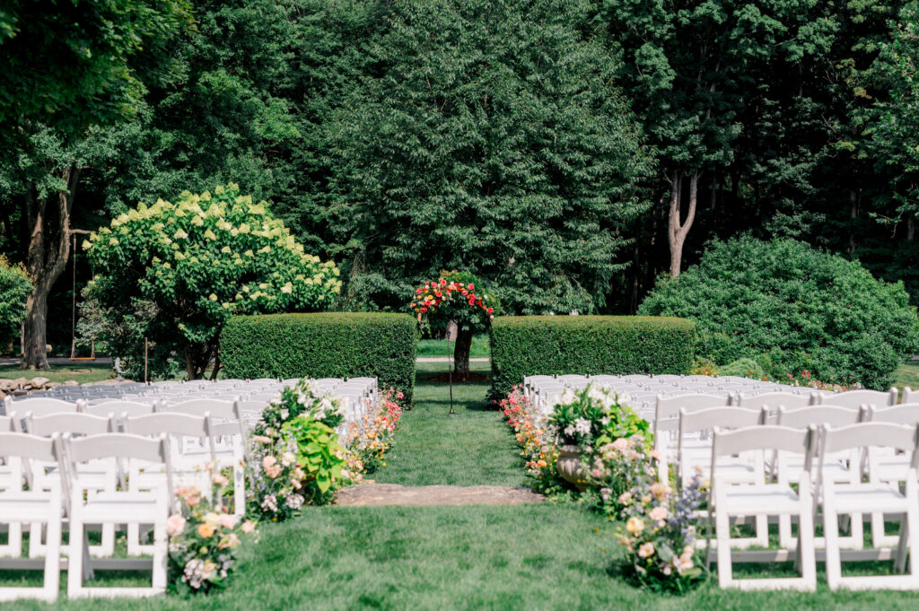 An outdoor ceremony setup at Smith Farm Gardens featuring rows of white chairs, vibrant floral arrangements, and a greenery-lined arch.