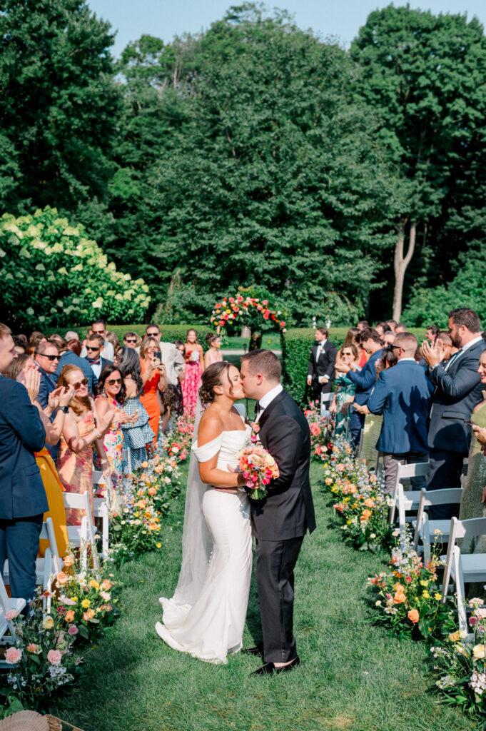 Bride and groom share a kiss at the end of the ceremony aisle at Smith Farm Gardens, surrounded by an applauding crowd.