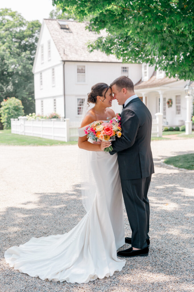 Bride and groom share an intimate moment under the shade of a tree with the charming white farmhouse of Smith Farm Gardens, one of the best wedding venues in Connecticut, in the background.