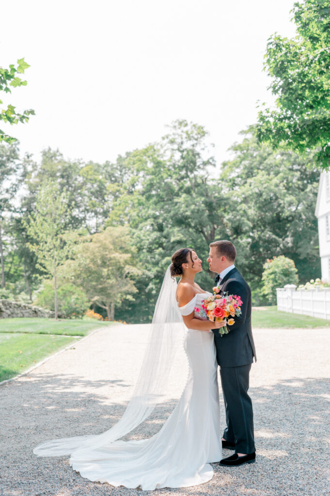 Bride and groom pose on a gravel pathway, with the bride holding a bouquet of vibrant flowers and the iconic Smith Farm Gardens estate behind them.