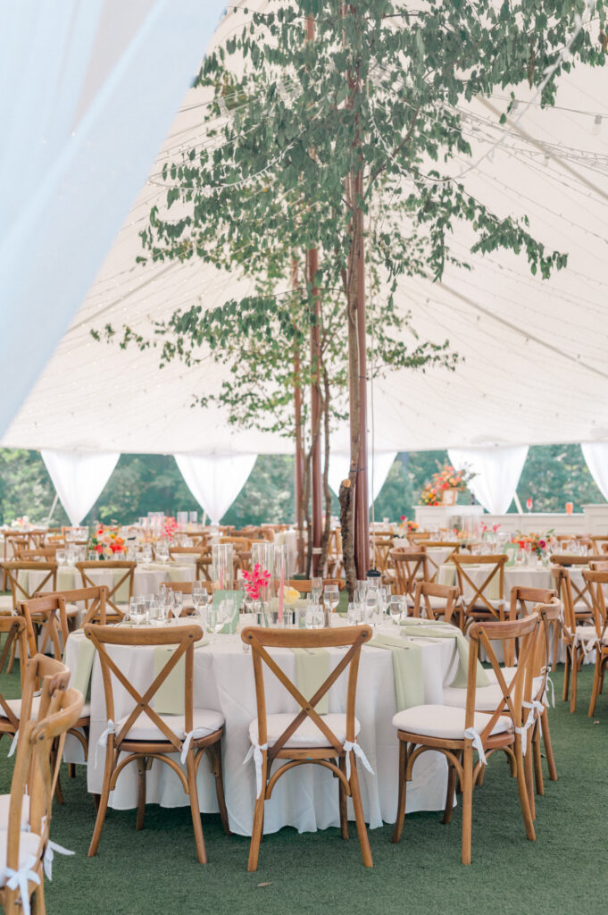 An elegantly decorated wedding reception under a sailcloth tent at Smith Farm Gardens, featuring round tables with wooden cross-back chairs, soft green accents, and floral centerpieces under a canopy of twinkling lights and greenery.