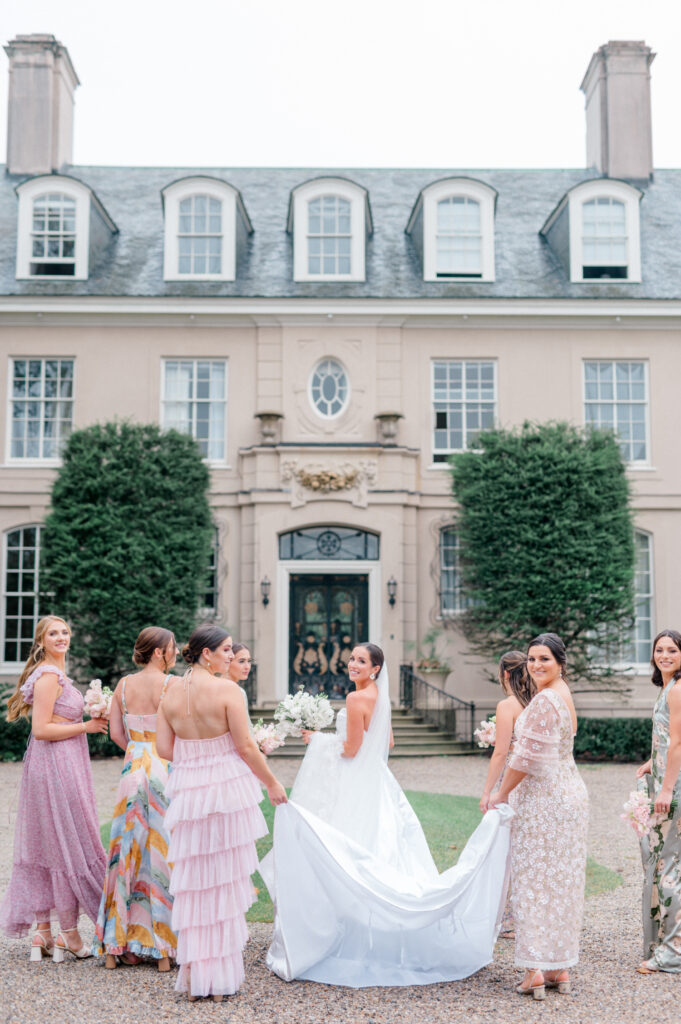 A bride surrounded by her bridesmaids, each dressed in pastel and floral dresses, standing in front of an estate with arched windows and lush greenery.