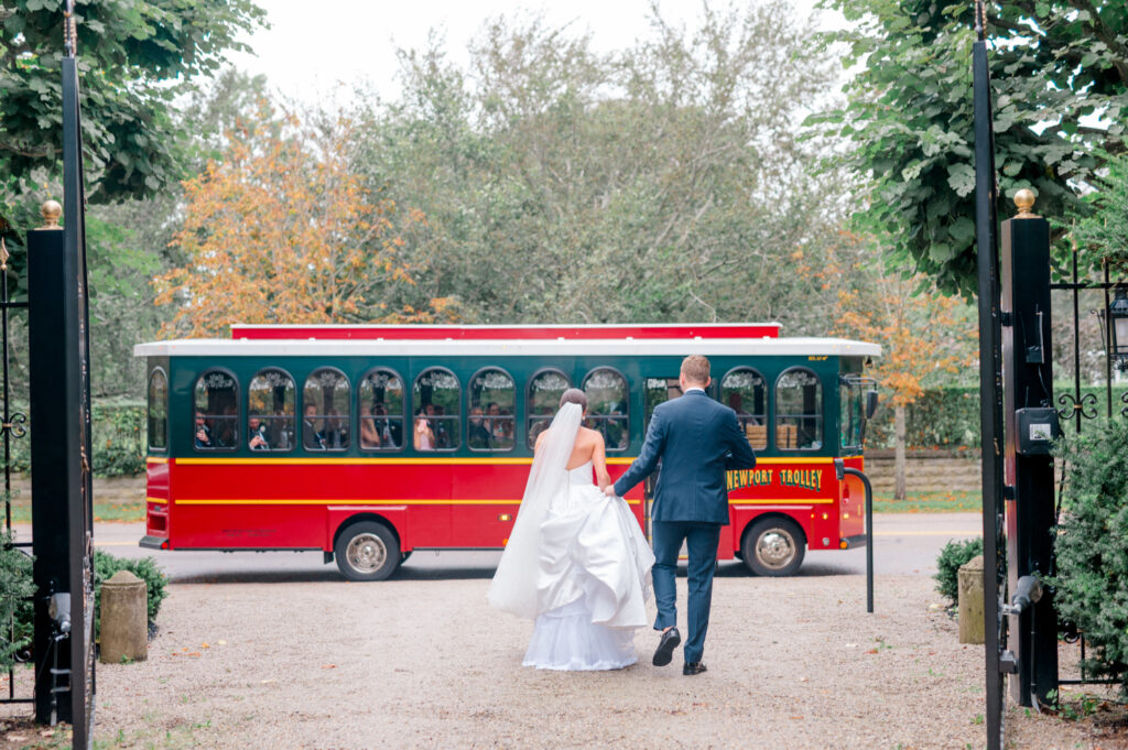 A bride and groom, hand in hand, walk toward a vintage red trolley surrounded by autumn trees and garden gates.