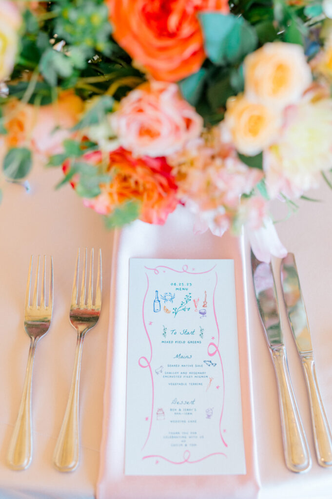 A close-up of a wedding menu on a neatly arranged place setting, featuring vibrant floral accents and polished silverware.