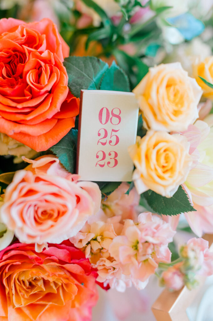 A close-up of bright orange, pink, and yellow flowers, highlighting a small card with the wedding date, "08.25.23."