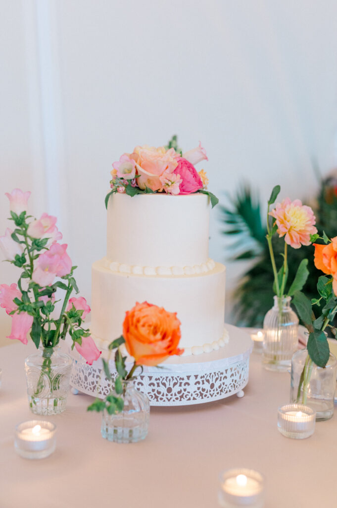 A simple two-tiered white wedding cake adorned with vibrant pink and orange blooms, surrounded bud vases of fresh flowers.