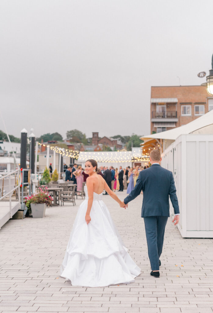 A bride and groom walk hand in hand along waterfront, with the bride glancing back with a big smile. The setting features warm string lights, mingling guests in the background.