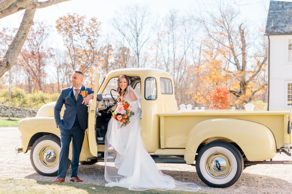 Bride and groom pose with a vintage yellow truck on a sunny autumn day. The bride is holding a vibrant bouquet of orange and pink flowers, while the groom leans against the truck in a navy suit with a matching boutonniere.