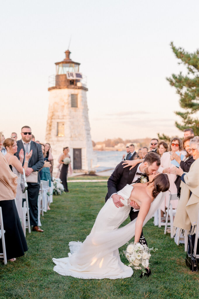 A groom dipping his bride into a kiss at the end of their outdoor ceremony aisle, with a scenic lighthouse and ocean in the background under soft golden hour light, perfectly illustrating a light and airy style of wedding photography.