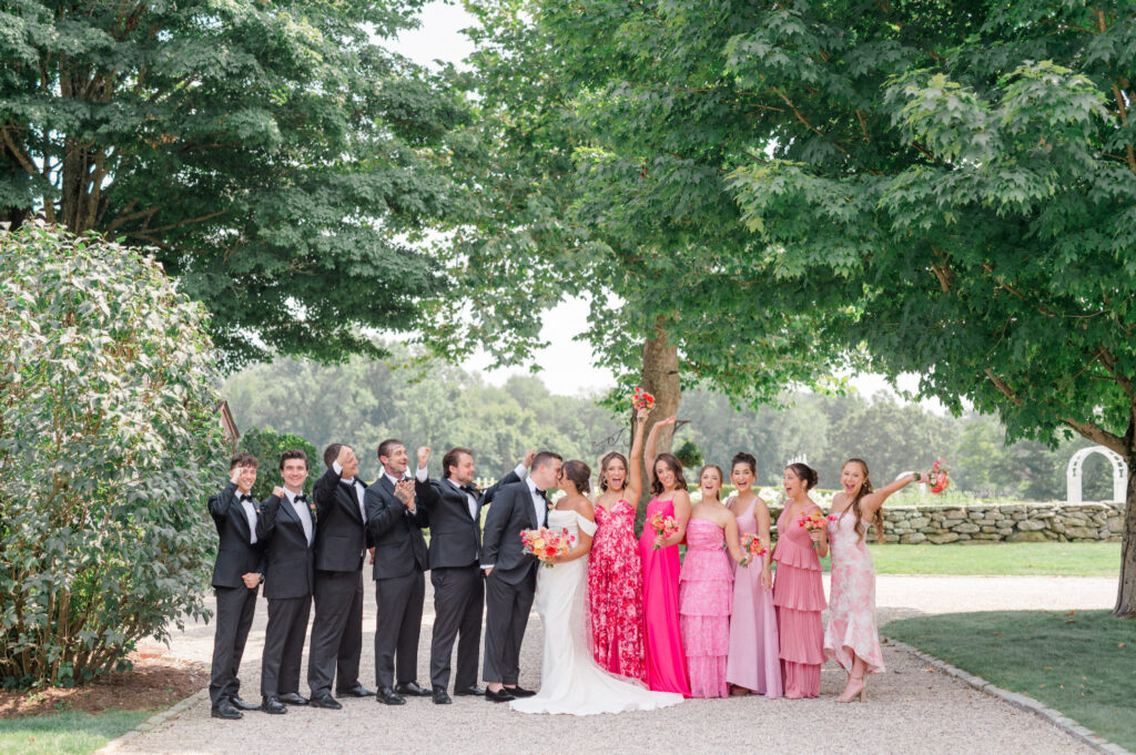 A wedding party celebrates outdoors under lush green trees, with bridesmaids dressed in vibrant pink dresses and groomsmen in classic black tuxedos. The bride and groom share a joyful moment in the center, surrounded by smiles and cheers in a garden setting.