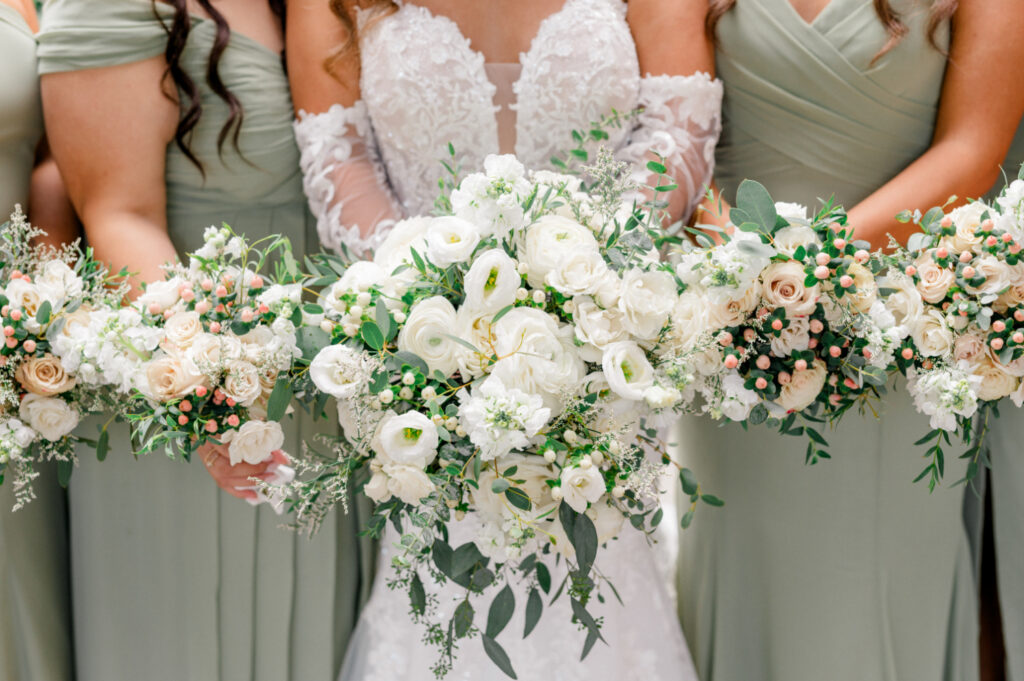 A close-up of the bride’s bouquet featuring lush white flowers, greenery, and subtle peach accents, held alongside matching bouquets carried by the bridesmaids in sage green dresses.