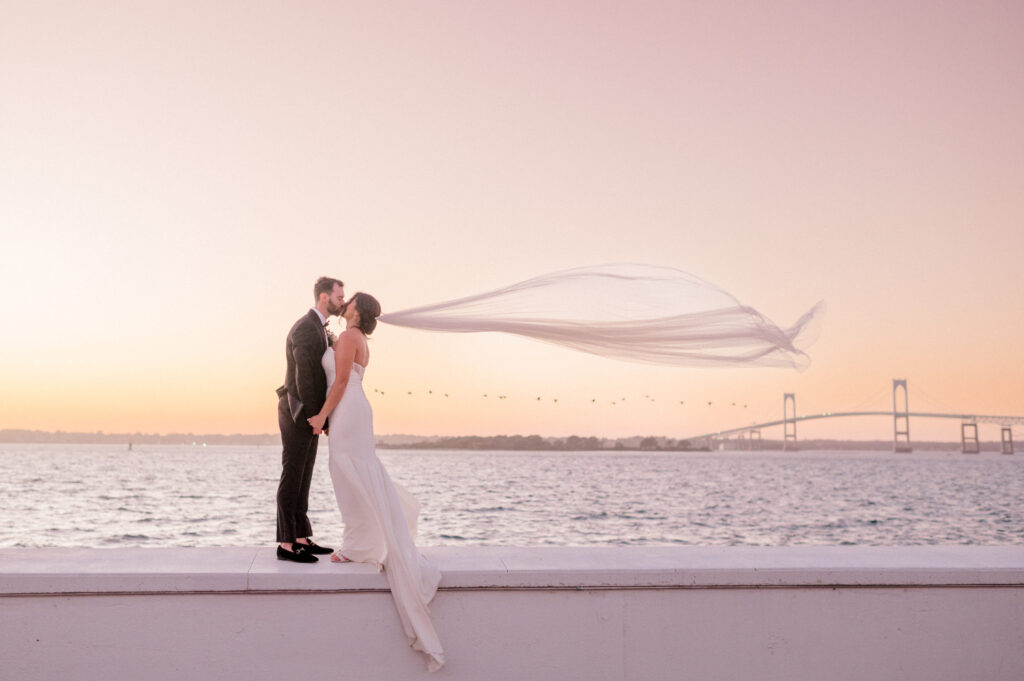 A bride and groom sharing a kiss as the bride’s veil flows dramatically in the wind against a waterfront sunset and a distant bridge in the background.