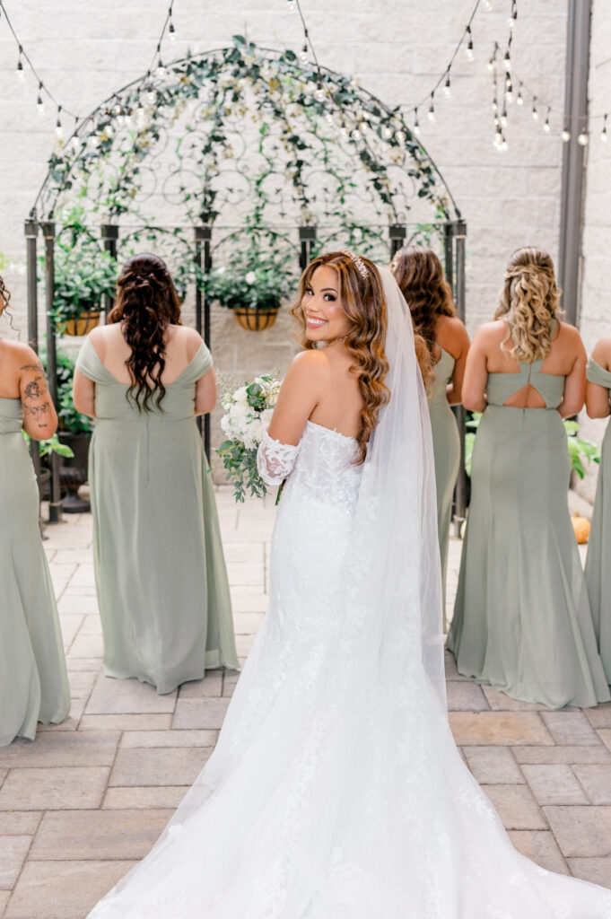 A bride in a fitted lace wedding gown stands smiling over her shoulder, holding a white floral bouquet with greenery. She is surrounded by her bridesmaids in sage green dresses, standing in front of a garden arch adorned with twinkling string lights.