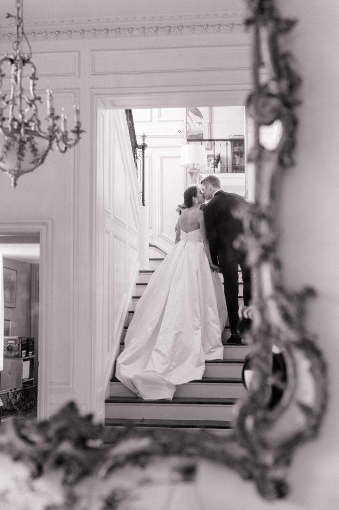 An elegant black-and-white photograph of a bride and groom sharing a romantic kiss on a grand staircase, reflected in an ornate mirror frame, perfectly reflecting an editorial style of wedding photography.