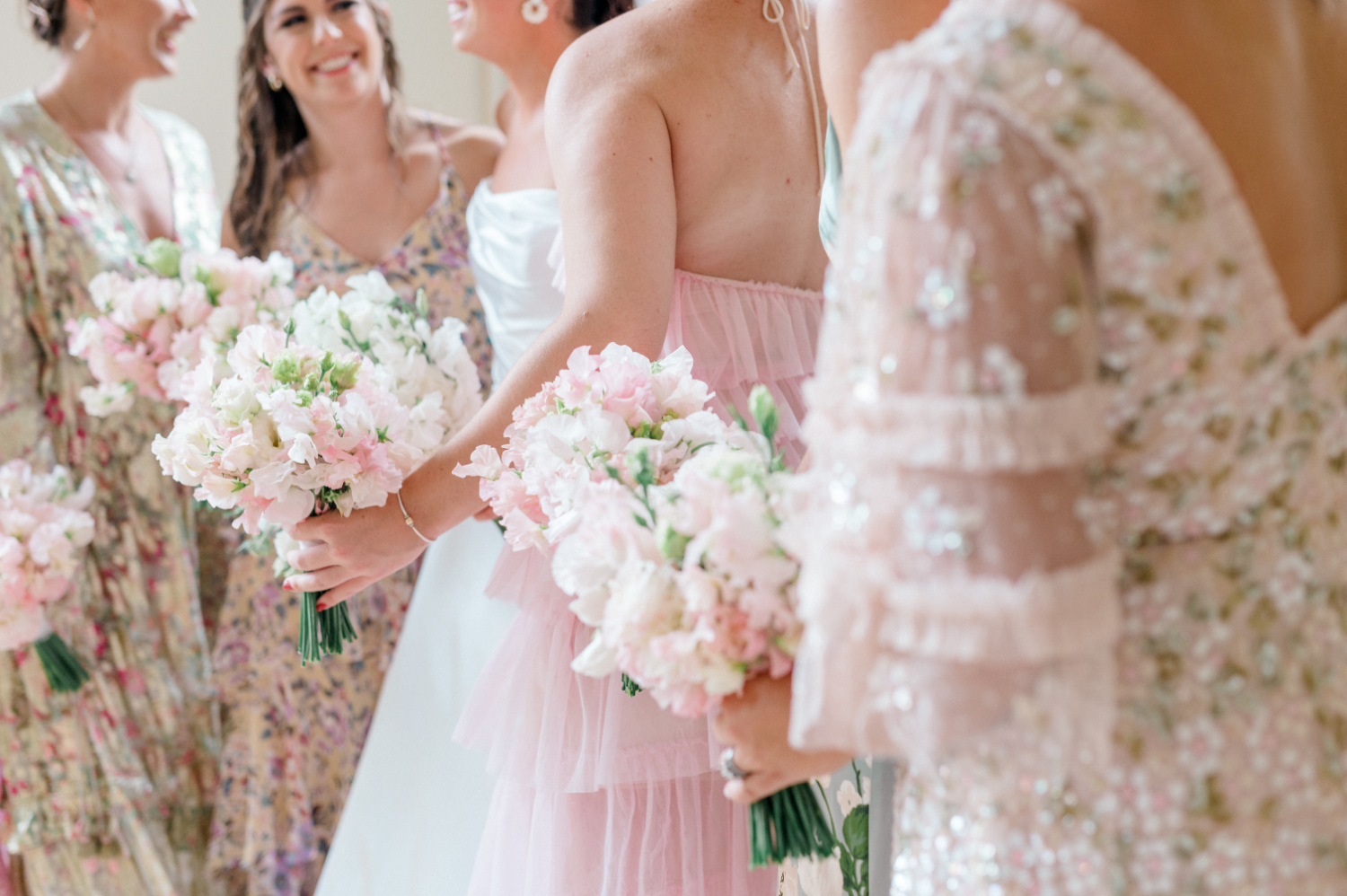 Bridesmaids and bride smiling and holding pastel pink and white bouquets, dressed in romantic, floral, and sequined dresses, showcasing a light and airy style of wedding photography.