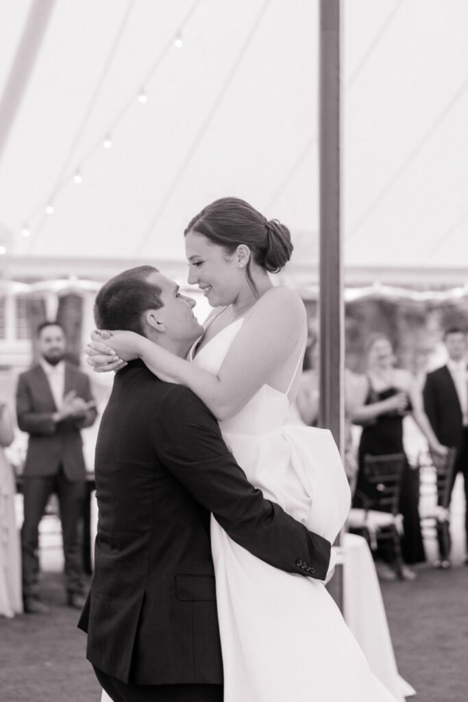 Black-and-white photo of the groom lifting the bride joyfully during their first dance, both smiling and surrounded by cheering guests.