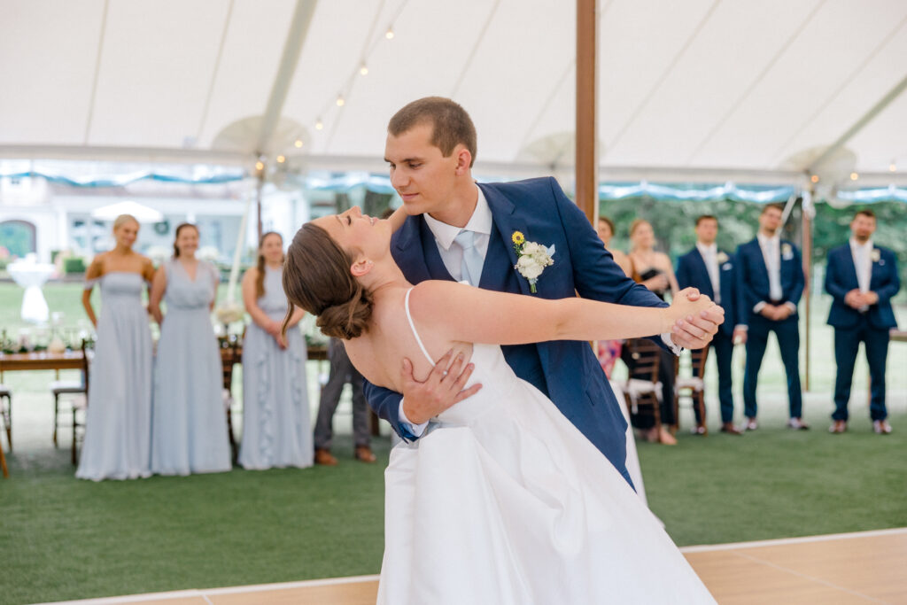 Groom dips the bride on the dance floor under a white sailcloth tent during their choreographed first dance at Smith Farm Gardens, with their wedding party smiling in the background.