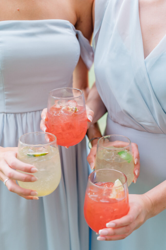 Bridesmaids toast with colorful signature drinks during cocktail hour, wearing elegant mismatched blue gowns at this summer wedding.