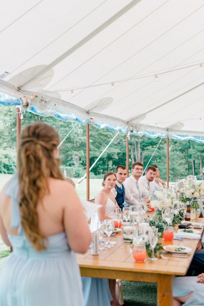 The bride and groom look on during a heartfelt toast given by a bridesmaid, surrounded by guests and floral details.
