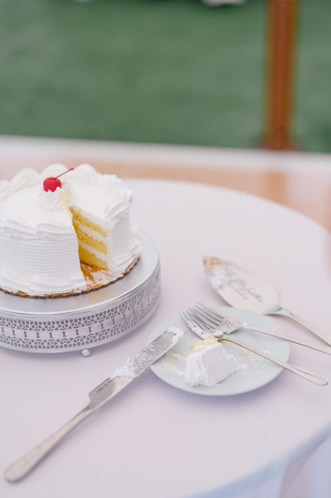 A simple, classic white wedding cake with a cherry garnish, partially sliced, sitting on a lace-trimmed cake stand.